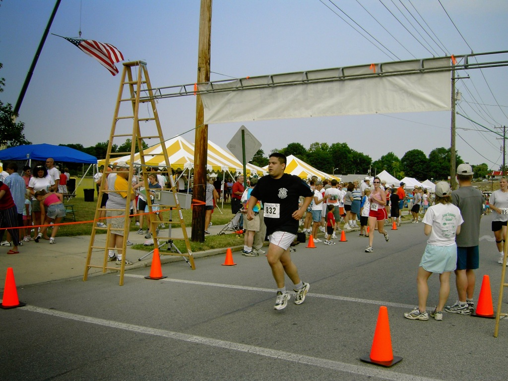 Image of a man crossing a finish line at a running event, wearing white shorts and a black tee shirt.