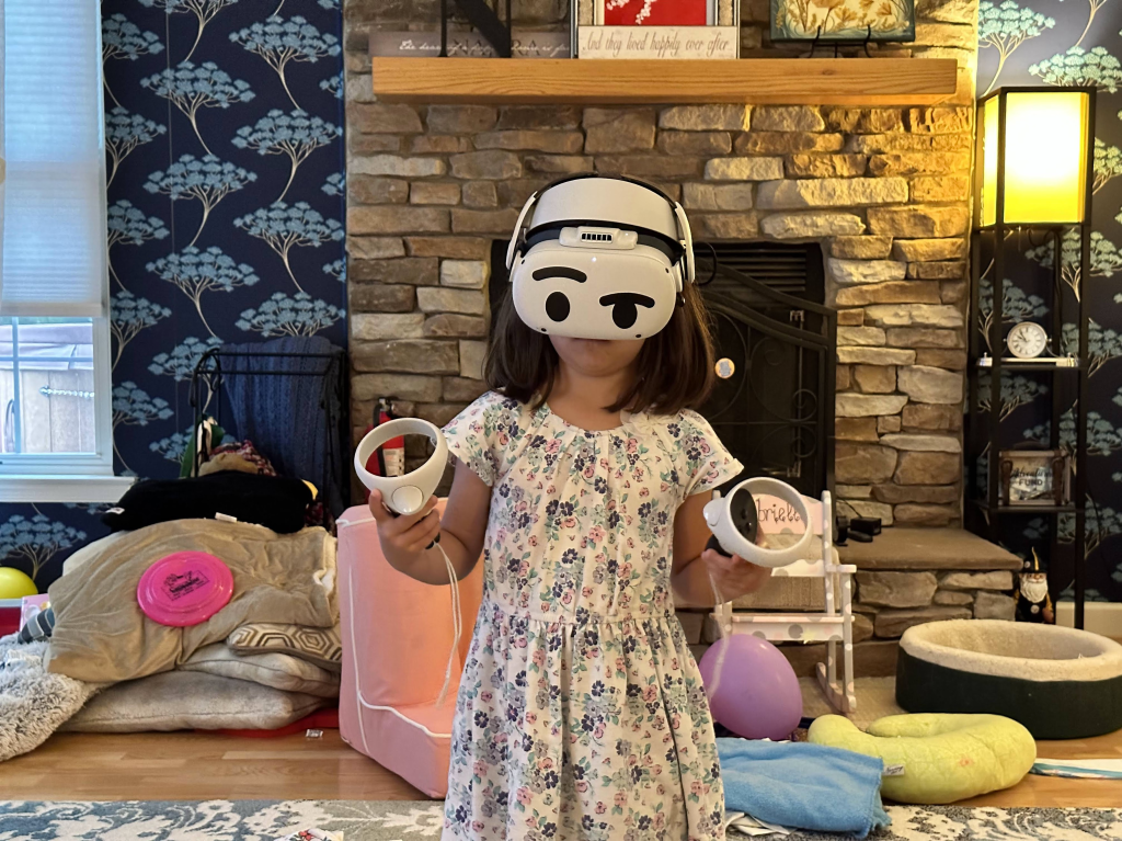 A little girl is wearing a virtual reality headset and grabbing two controllers. She is in a living room with cushions and toys on the floor behind her.